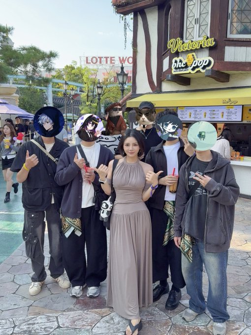 A group photo taken at Lotte World, showing a woman in a beige dress posing with several people whose faces are covered by various character masks or avatars.