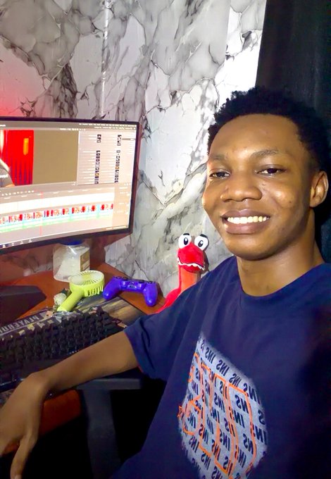 A young man smiling at a desk with a video editing software open on his monitor, with an Elmo plush toy, a keyboard, and gaming controllers visible in the background.