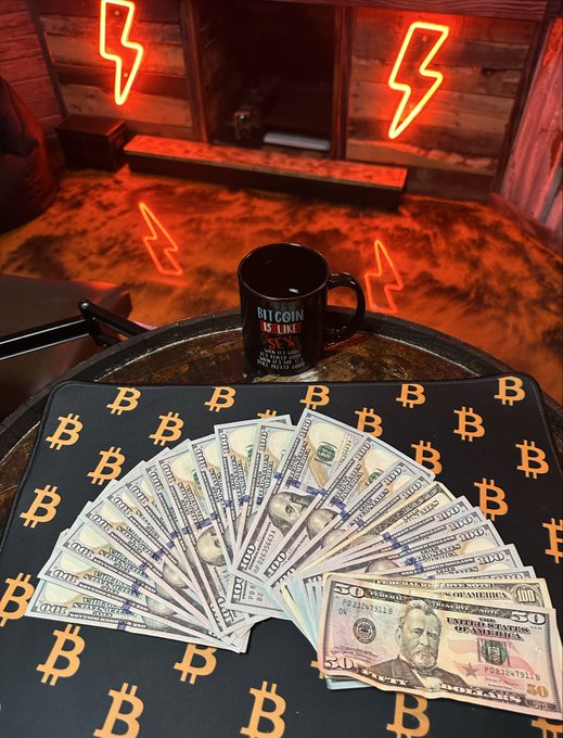 A fan of US $50 and $100 bills spread on a Bitcoin-symbol desk mat, with a 'Bitcoin is Like Sex' mug and neon lightning bolt signs in the background.