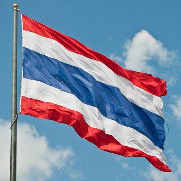 The national flag of Thailand, featuring horizontal red, white, and blue stripes, waving against a partly cloudy sky.