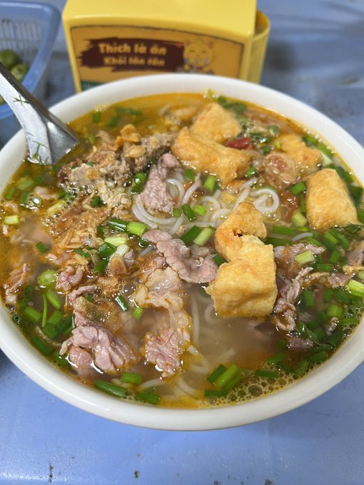 A close-up photograph of a Vietnamese noodle soup, likely bún bò Huế, featuring rice noodles, sliced beef, fried tofu cubes, and green onions in a rich broth, served in a white bowl with a spoon, with a yellow container labeled "Thịt lợn Khô Hòa Bình" visible in the background.