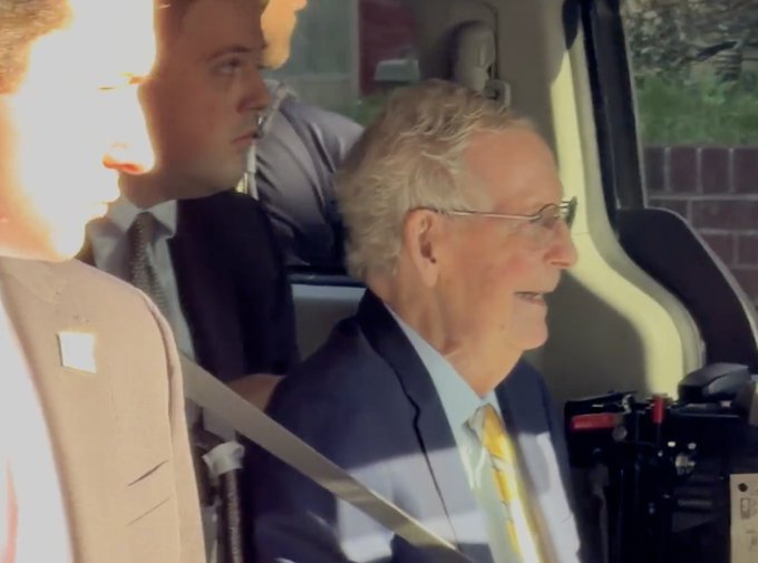 A photograph of an elderly man wearing glasses and a suit with a yellow tie, sitting in the back seat of a vehicle with a seatbelt on, accompanied by other passengers.