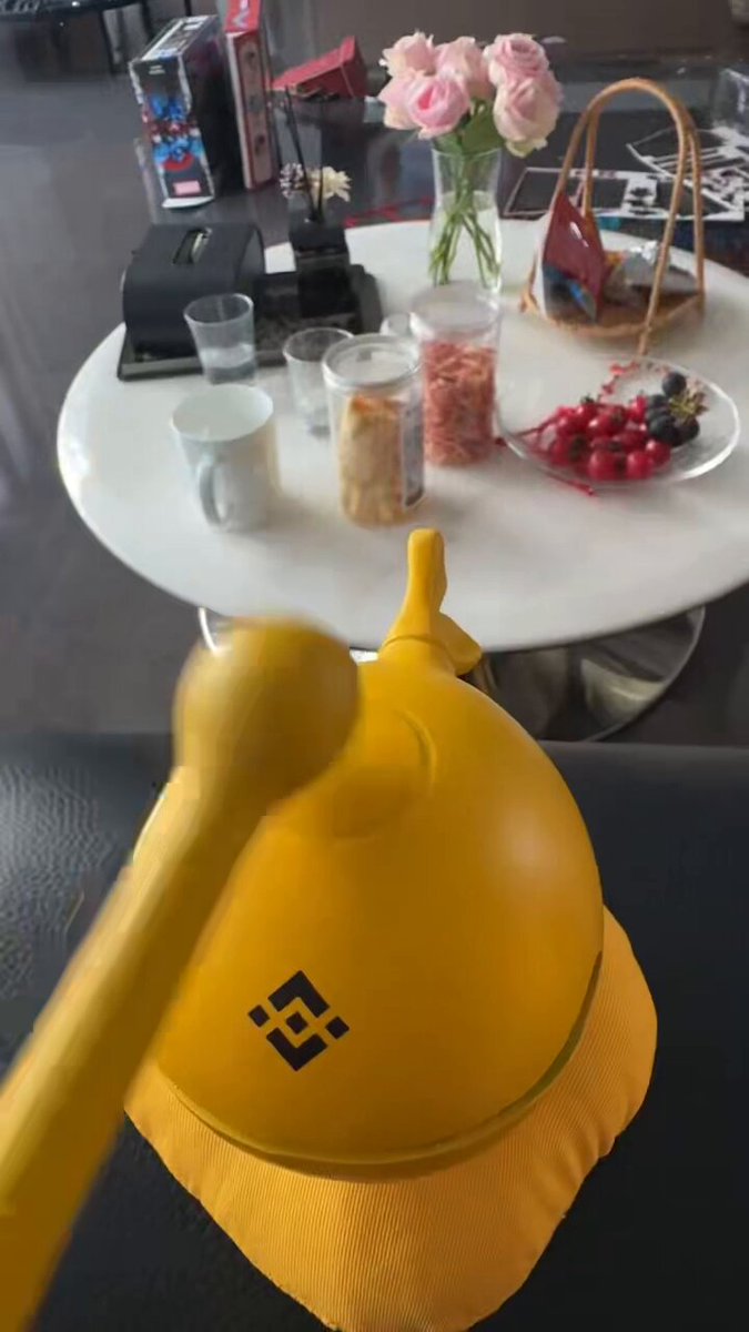 The image shows a yellow object, possibly a hat or a lamp, sitting on top of an item such as a lampshade, tablecloth, or cushion. There are several other items present, including decorative vases, fruit like apples, oranges, and strawberries, cups, glasses, a bowl, and plates with various foods arranged around the yellow object. The image also appears to be connected to a post on social media.