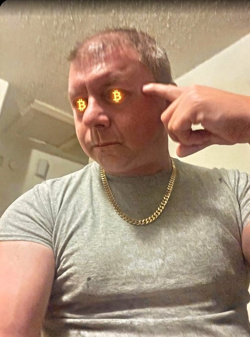A man with a gold chain points to his temple with glowing Bitcoin symbols overlaid on his eyes.