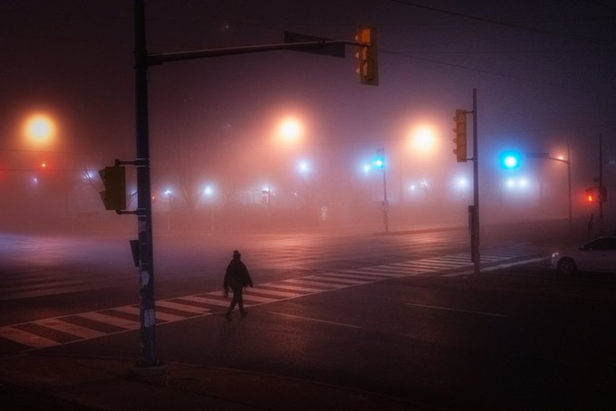 A foggy nighttime urban intersection with glowing streetlights and traffic signals in red and blue, where a solitary figure in dark clothing walks across the crosswalk.
