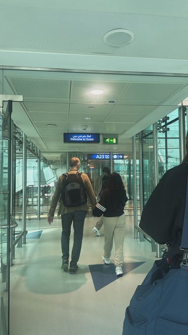 A couple walks through an airport terminal, following signs for 'Welcome to Dubai' in Arabic and English, with directional signs pointing to gates A23.