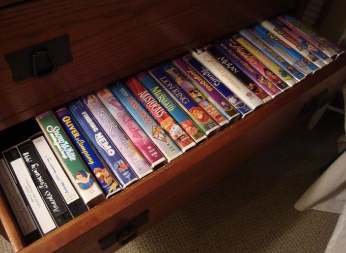 An open wooden drawer filled with a collection of VHS tapes and Disney VHS movies, including visible titles like Aladdin, The Lion King, Cinderella, The Little Mermaid, Mulan, and Oliver & Company.