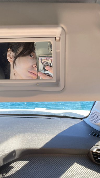 A woman takes a mirror selfie in a car, her reflection captured in the sun visor mirror with the ocean visible through the windshield below.