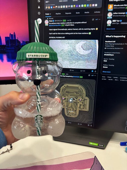 A hand holds a clear bear-shaped Starbucks tumbler with a green lid and a green-and-white striped straw in front of a laptop screen showing Twitter/X posts, including one about Hozier and the album "Unreal Unearth."