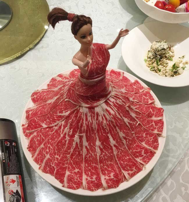 A creative presentation of thinly sliced raw beef arranged in a circular skirt pattern around a doll figurine, with side dishes visible on the table.
