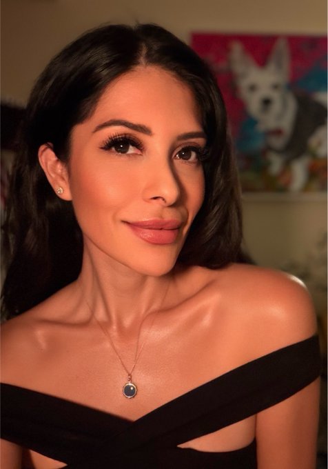 A woman with dark hair and a polished, glamorous look smiles at the camera while wearing a black off-the-shoulder top and a delicate pendant necklace, embodying self-confidence and self-care—perfectly complementing the message "Invest in yourself. It pays the best interest." In the background, a colorful painting of a white dog adds a personal, artistic touch to the elegant portrait.