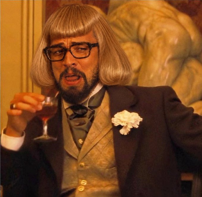 A man with a beard, glasses, and a gray bob wig wearing a formal suit with a white carnation and holding a glass of wine.