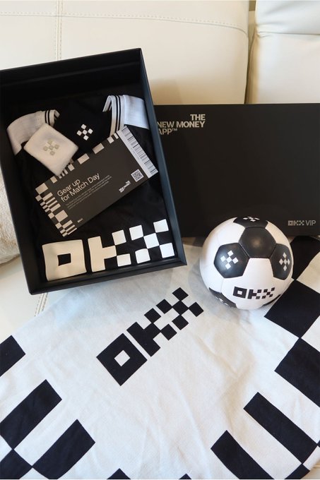 An OKX merchandise box containing a black and white 'Geor Up for Match Day' jersey and socks, displayed alongside an OKX soccer ball and matching towel.