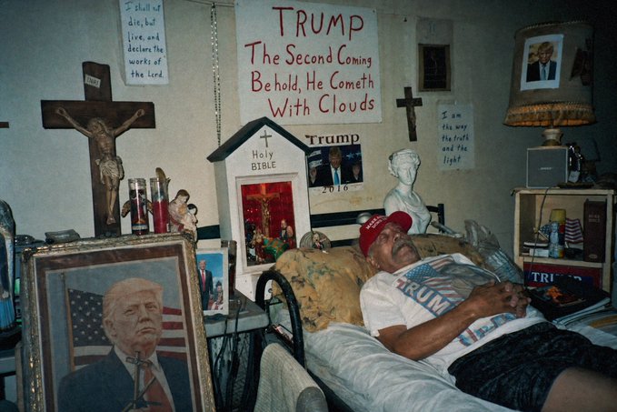 A man wearing a 'TRUMP' shirt and a red MAGA hat rests in a bed surrounded by religious and political imagery, including signs reading 'TRUMP The Second Coming Behold, He Cometh With Clouds' and 'I shall not die, but live, and declare the works of the Lord.'