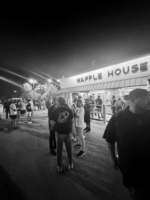 A black-and-white photograph captures a crowd of people gathered outside a brightly lit 'WAFFLE HOUSE' restaurant at night.