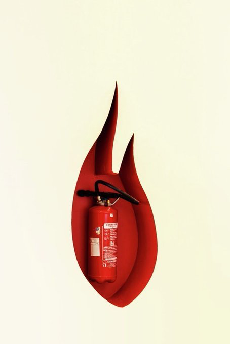A red flame-shaped decorative cover encases a fire extinguisher mounted on a cream-colored wall, with visible warning labels and instructional text on the canister.