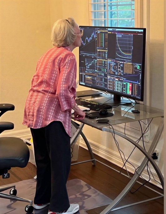 A woman stands focused and riveted, leaning closely toward a large monitor displaying multiple trading charts and graphs in a home office setup. Her intense concentration on the market data embodies the caption's message about being completely "locked in" while watching the markets.