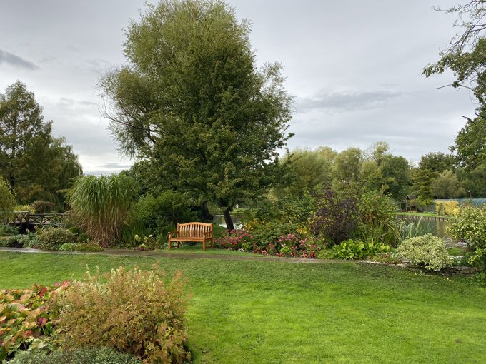 A tranquil garden landscape featuring a wooden bench nestled among lush greenery, vibrant flowers, and a large tree under a cloudy sky.