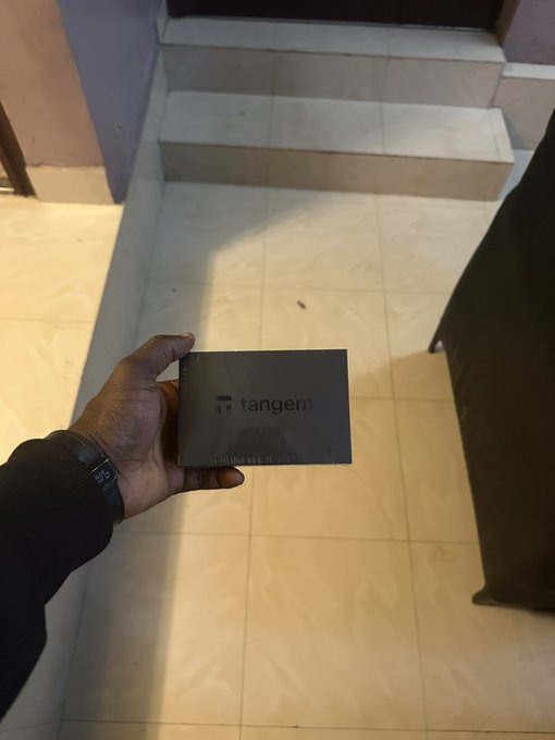 A person's hand holds a black envelope labeled 'tangem' indoors near a staircase, with the person wearing a black sleeve and wristband.