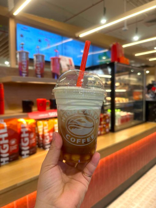 A hand holds a clear plastic cup of iced coffee from Highlands Coffee in front of a store counter displaying red product packaging.
