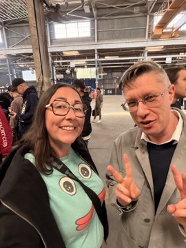 A smiling woman in a cartoon-eyed shirt and a man in a gray blazer pose with peace signs inside a busy industrial hall.