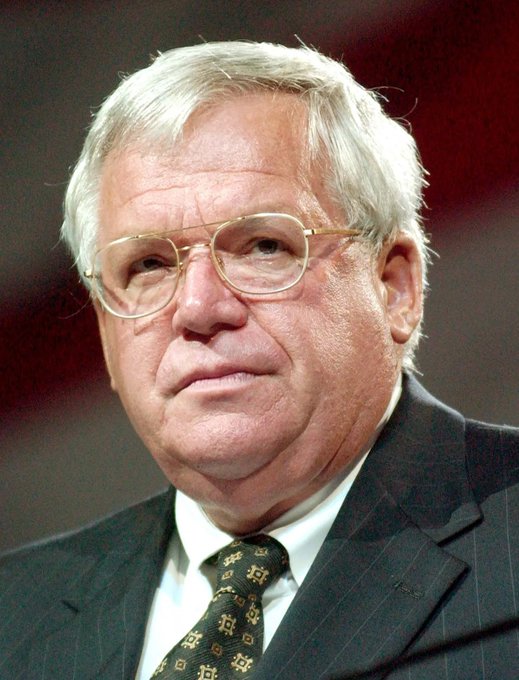 An older man with white hair, wearing wire-rimmed glasses, a dark pinstripe suit, and a patterned tie, looks off to the side with a serious expression. The image accompanies the news of Dennis Hastert's passing, who served as the longest-tenured Republican Speaker of the House.