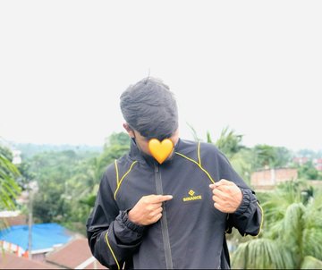 A man wearing a black and yellow jacket zips up his jacket with a heart emoji covering his face, set against a backdrop of trees and rooftops.
