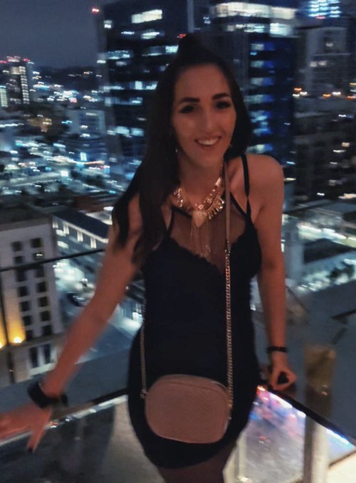A woman in a black dress wearing a statement necklace and carrying a small crossbody bag smiles at the camera, posed against a backdrop of city lights visible from a high-rise balcony.
