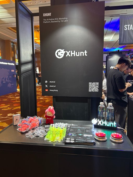 An XHUNT exhibition booth at a Hong Kong event, described as 'The AI-Native KOL Marketing Platform, Backed by Yzi Labs,' displays promotional items including water bottles, branded merchandise, and a pink mascot plush.