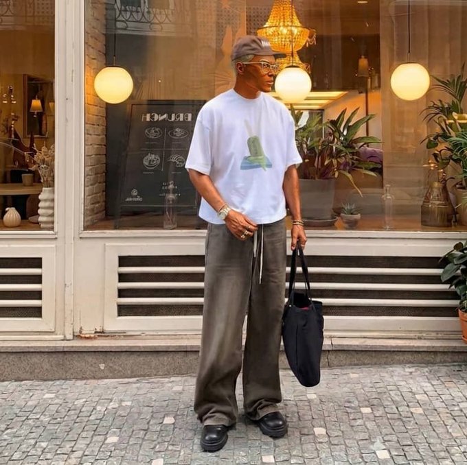 A man wearing a white graphic t-shirt, brown wide-leg pants, a cap, and glasses stands on a cobblestone street holding a black tote bag, with a cafe interior visible behind him.