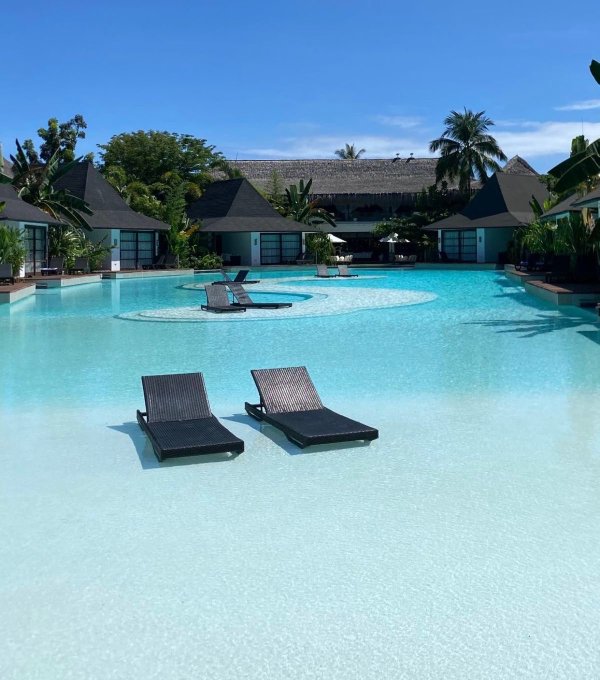 A luxurious tropical resort with a large turquoise swimming pool, black lounge chairs floating in the water, thatched-roof buildings in the background, and palm trees under a clear blue sky.