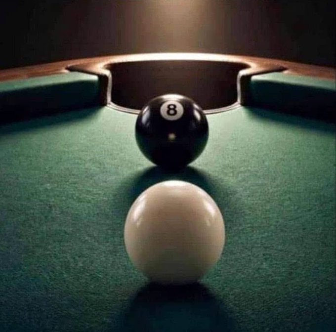 A pool table scene with a white cue ball positioned in the foreground facing a black 8-ball near the corner pocket, lit by an overhead spotlight.
