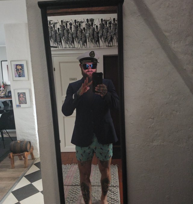 A mirror selfie of a man with a beard wearing a dark blazer, light blue patterned shorts, a captain's hat, and reflective sunglasses, with artwork visible on the walls around the mirror.