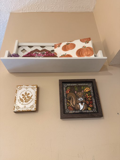 An autumn-themed decorative display on a shelf featuring a wooden squirrel stained-glass-style plaque, a 'Beginnings' seed packet, and a white tray with 'Hello Fall' pumpkin decor.