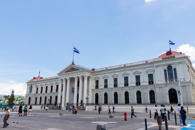 A grand neoclassical government building with white columns and a flag flying above its pediment stands under a bright blue sky, with people walking across the plaza in front. The imposing architecture symbolizes official state authority, reinforcing the post's message about government efforts to regulate and oversee crypto activities like Bitcoin.