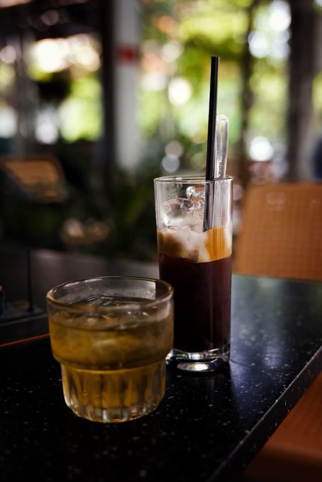 Two beverages sit on a dark table at an outdoor café: a short glass with an amber-colored iced drink and a tall glass containing a layered iced coffee with a black straw.