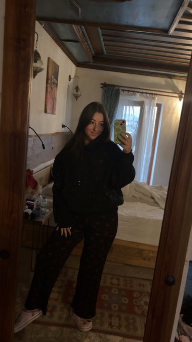 A young woman takes a mirror selfie in a cozy bedroom with wooden ceiling beams, wearing a black sweater and dark patterned pants while holding her phone.