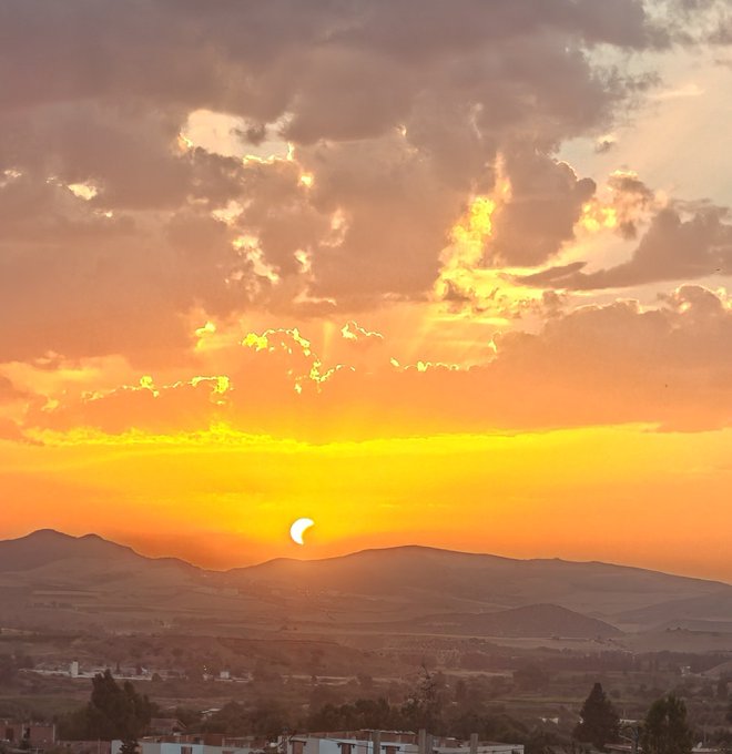 A scenic photograph of a partial solar eclipse at sunset, with the sun visible above a silhouette of mountains and clouds reflecting warm colors.