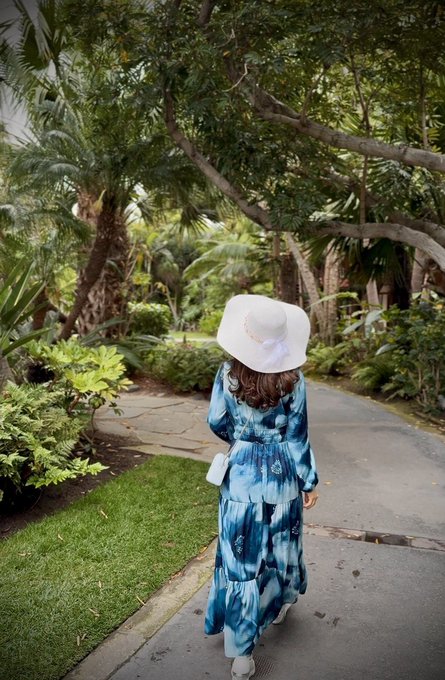 A woman wearing a blue tie-dye tiered dress and white sun hat walks along a garden path surrounded by lush tropical plants and trees.