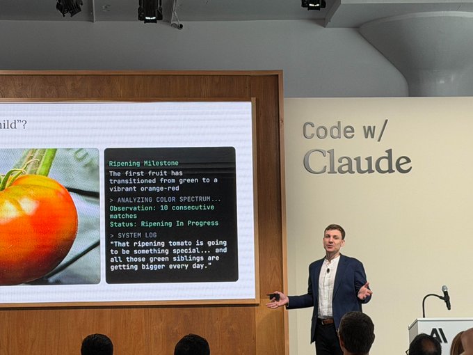 The image shows a man standing behind a podium at an event, giving a presentation or speech. The room has white walls and a wooden floor, creating a professional atmosphere for the gathering. On the screen behind him, there's a slide with text that reads "Code/Claude" and "Ripping Stone". This suggests that the image might be related to a project or an event involving code or programming.