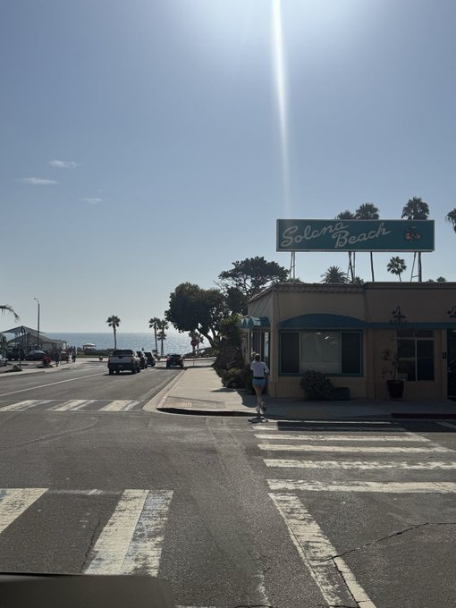 A coastal street scene features a teal "Solana Beach" sign atop a building near the ocean, with palm trees lining the sunny boardwalk area. The image perfectly captures the laid-back "Solana summer" vibe mentioned in the caption, showcasing the sunny weather and beachside charm of this Southern California town.