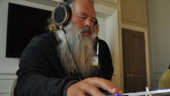 An older man with a long gray beard wearing headphones, focused while working at a keyboard or mixing board.