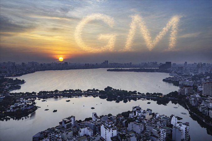 An aerial view of a city skyline at sunset over a lake, with the letters 'GM' (good morning) creatively formed by clouds in the sky.