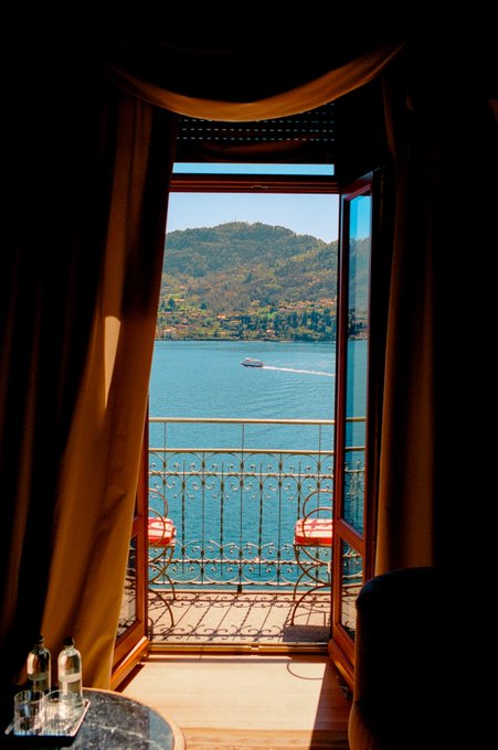 A scenic view looking out through an open window framed by brown curtains, revealing a balcony with chairs overlooking a bright blue lake, forested hills, and a passing boat.
