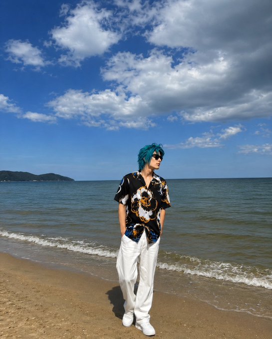 A person with teal hair wearing sunglasses, a black patterned shirt, and white pants standing on a sandy beach with the ocean and cloudy sky in the background.