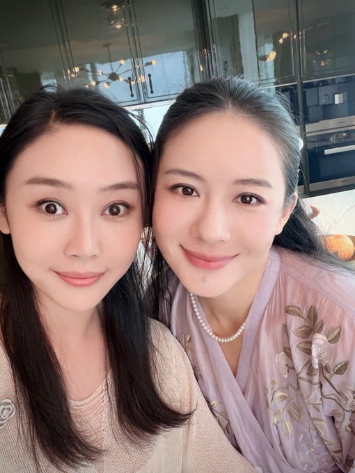 A selfie of two young women smiling together, one wearing a beige top and the other in a lavender embroidered top with a pearl necklace.