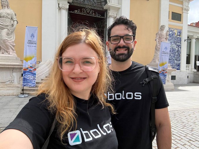 A selfie of two people wearing matching black 'bolos' t-shirts standing in front of a building with ETN LISBON banners, featuring ornate Portuguese tile work and statues.