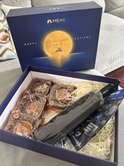 An open MEXC gift box labeled 'Happy Mid Autumn Festival 2020' displays mooncakes and other packaged items resting on shredded paper filler.