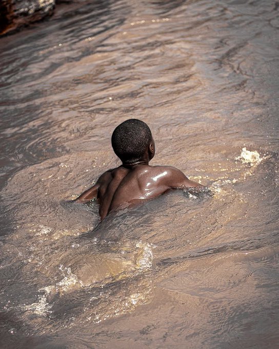 A young boy with short hair wades through murky brown water, his back and shoulders glistening wet.