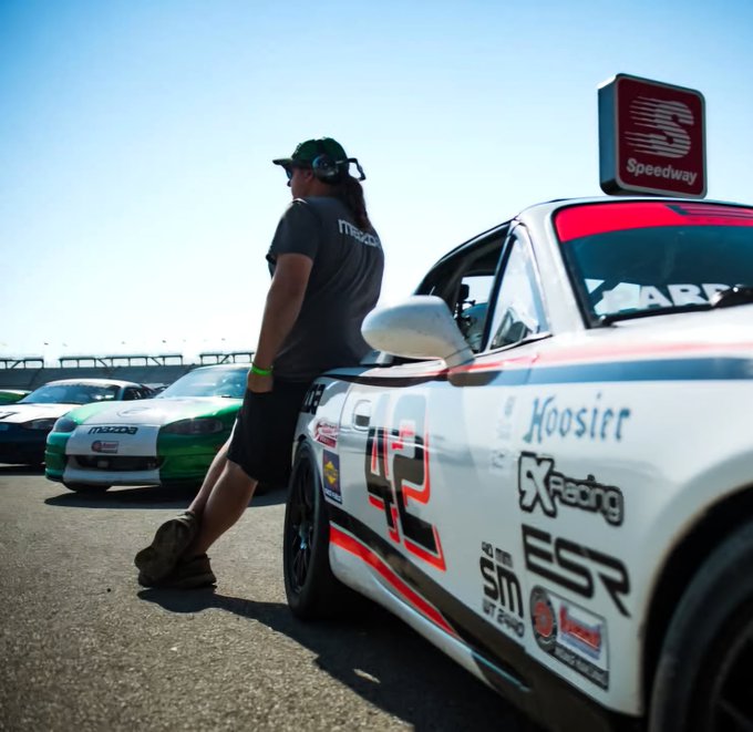 A person in a Mazda cap and Mazda-branded shirt leans against a white race car with Hoosier and SX Racing decals at a Speedway event, with other Mazda cars visible in the background.